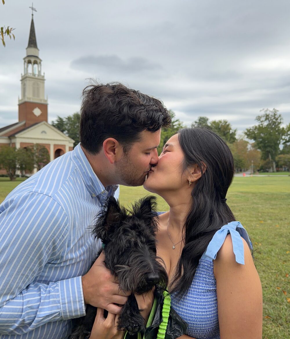 Joy and Michael kissing while holding their dog, a chapel behind them